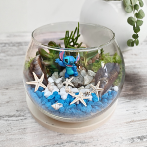 Ocean-themed glass terrarium with succulents, blue pebbles, seashells, starfish, moss, and a small figurine in a round glass bowl.