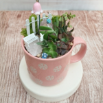 Succulent mini garden in a pink ceramic mug with moss, pebbles, and fairy garden décor including a white bench and tiny mushrooms.