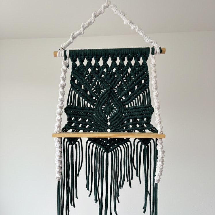 Macrame shelf Green macramé wall hanging with wooden shelf and fringe detailing, suitable for 9–12cm indoor plant pots, displayed against a white wall.