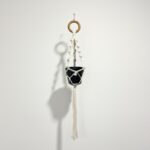 Short natural cotton macramé plant hanger with wooden ring holding a black 12cm nursery pot, hanging against a white wall.