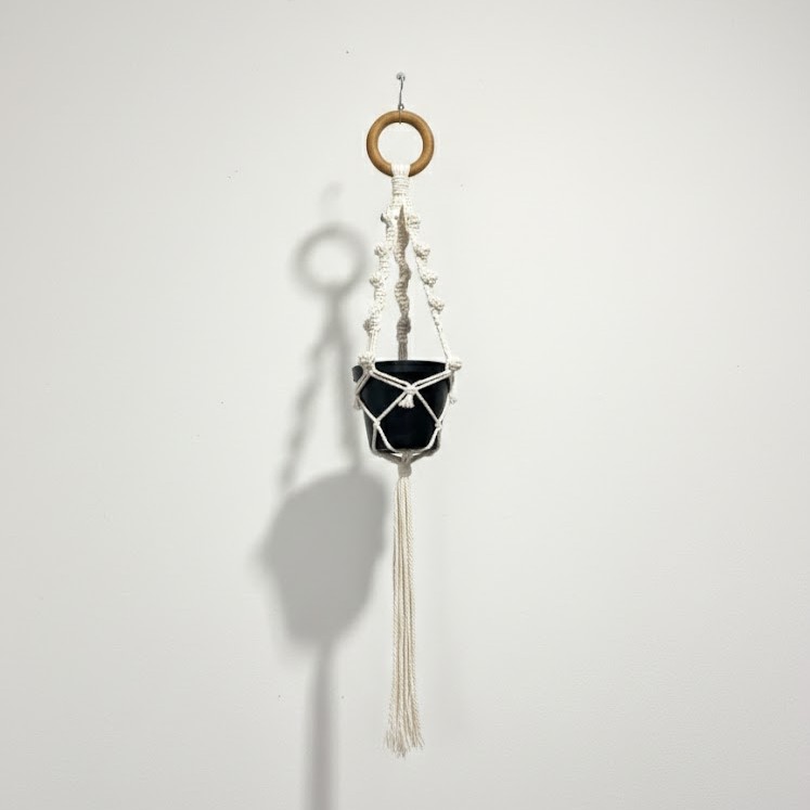 Macrame small basket Short natural cotton macramé plant hanger with wooden ring holding a black 12cm nursery pot, hanging against a white wall.